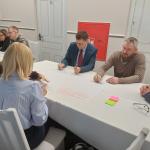 Sześć osób siedzi przy stole, piszą na dużym arkuszu papieru, notatki i kolorowe karteczki.