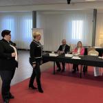 Dwie kobiety stoją przed stołem, gdzie siedzi czworo ludzi podczas spotkania.