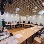 Osoby siedzące przy stołach na spotkaniu w jasnej sali konferencyjnej.